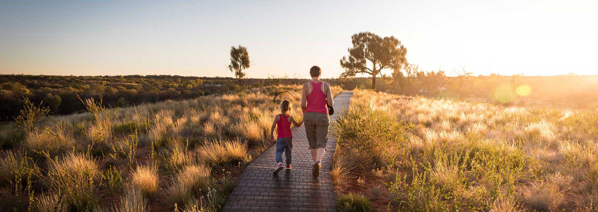 woman and girl wearing pink tank tops walking down paved pathway in large open field at sunset